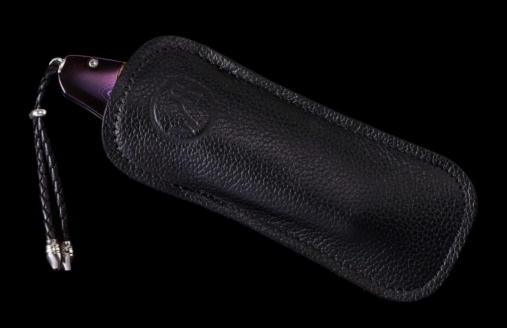Purple pattern-handled knife partially sheathed in a black pebbled leather sheath with an embossed logo and braided lanyard.