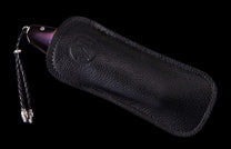 Purple pattern-handled knife partially sheathed in a black pebbled leather sheath with an embossed logo and braided lanyard.