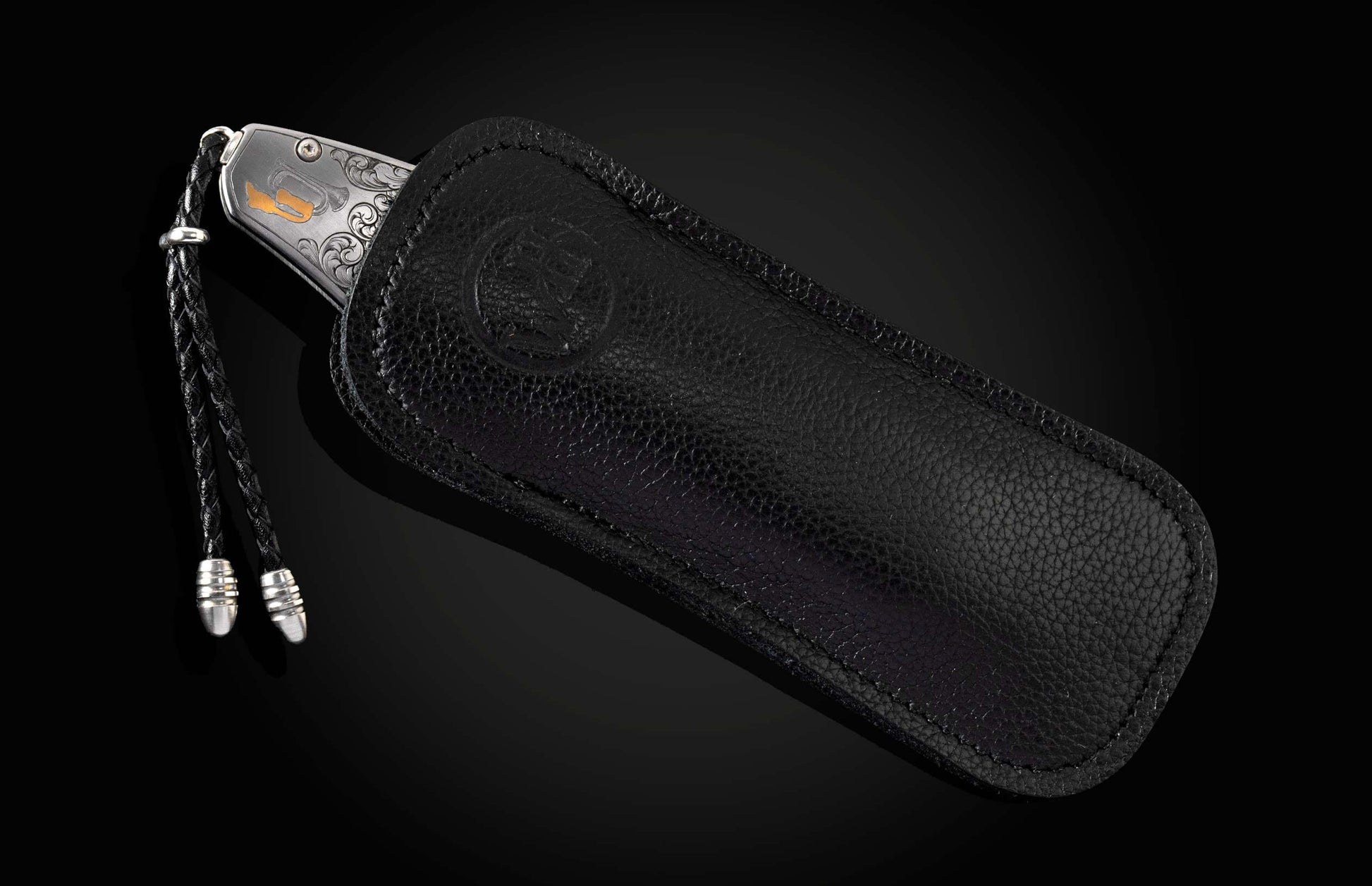 Engraved silver knife with gold horn detail, partially in a black textured leather sheath featuring an embossed logo and braided lanyard.