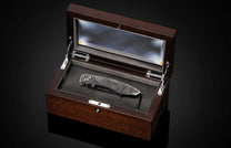 Dark folding knife with intricate scrollwork handle, displayed in an open dark wood box with glass top.