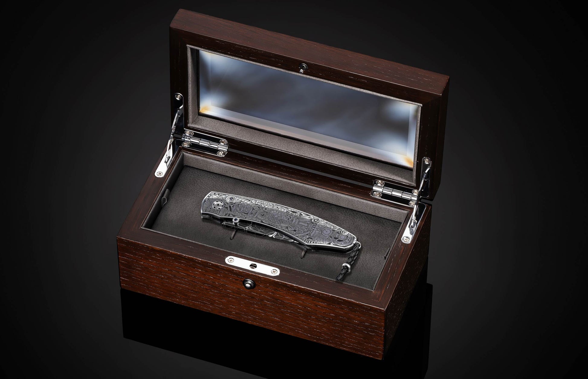 Silver-toned folding knife with intricate steampunk engraving resting in an open dark wood display box with a glass lid.
