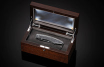 Silver-toned folding knife with intricate steampunk engraving resting in an open dark wood display box with a glass lid.