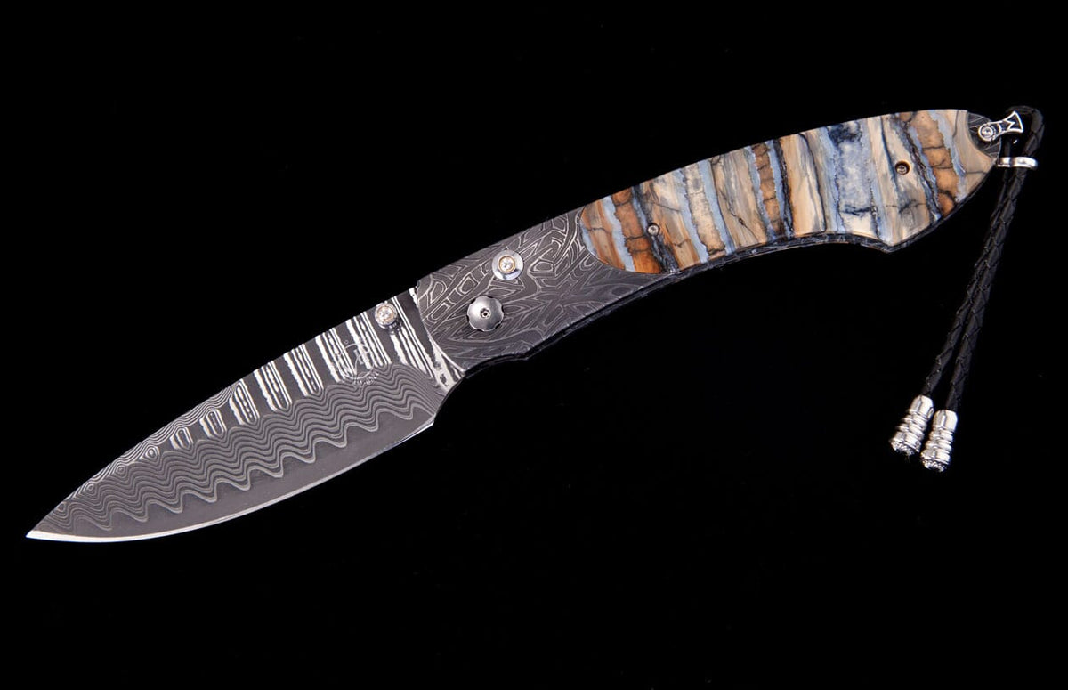 Damascus steel folding knife by Tamreen with a wavy blade, geometric handle, layered blue and brown scales, and a leather lanyard.