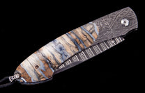 Closed folding knife with a multi-toned petrified wood handle by Tamreen, featuring intricate Damascus steel patterns and a black leather lanyard.