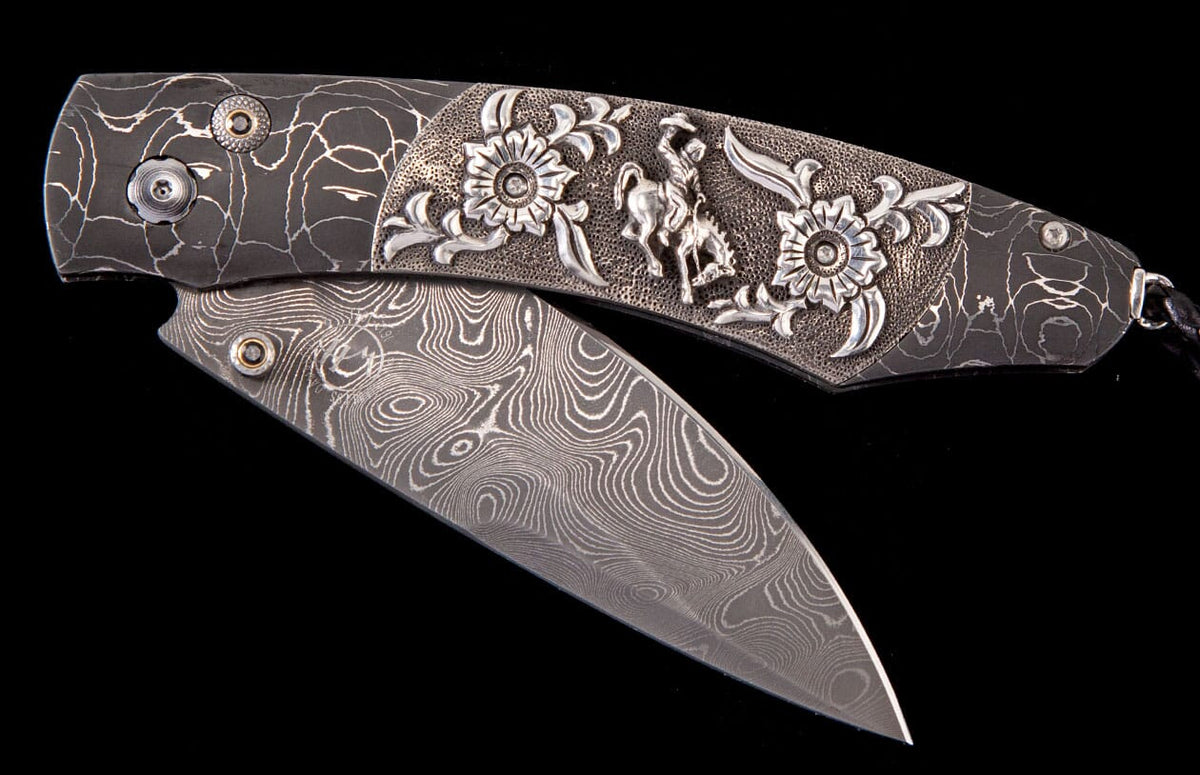Damascus steel folding knife with a dark patterned handle featuring silver cowboy and floral embellishments.