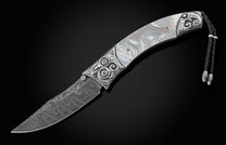 Handcrafted pocket knife from William Henry featuring a frame in hand-carved sterling silver inlaid with mother of pearl. The blade is hand-forged Damascus steel and the button lock and the thumb stud are set with white topaz gemstones. The knife is displ