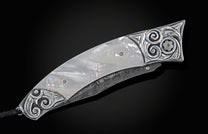 Handcrafted pocket knife from William Henry featuring a frame in hand-carved sterling silver inlaid with mother of pearl. The blade is hand-forged Damascus steel and the button lock and the thumb stud are set with white topaz gemstones. The knife is displ