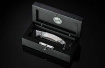Handcrafted pocket knife from William Henry featuring a frame in hand-carved sterling silver inlaid with mother of pearl. The blade is hand-forged Damascus steel and the button lock and the thumb stud are set with white topaz gemstones. The knife is displ