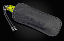 Knife with vibrant green and brown handle partially inserted into a black textured leather case with an embossed logo and braided lanyard.