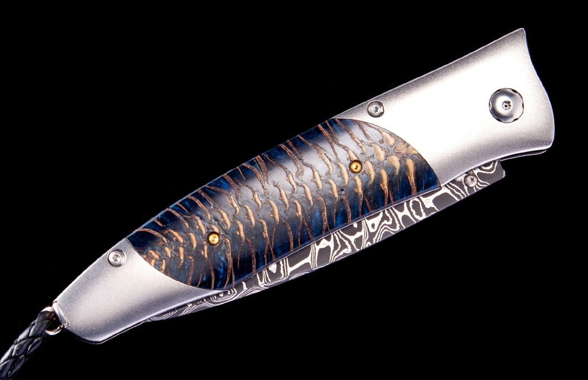 Silver-handled folding knife by Tamreen, featuring a blue fish scale-textured inlay, Damascus steel details, and a braided leather lanyard.