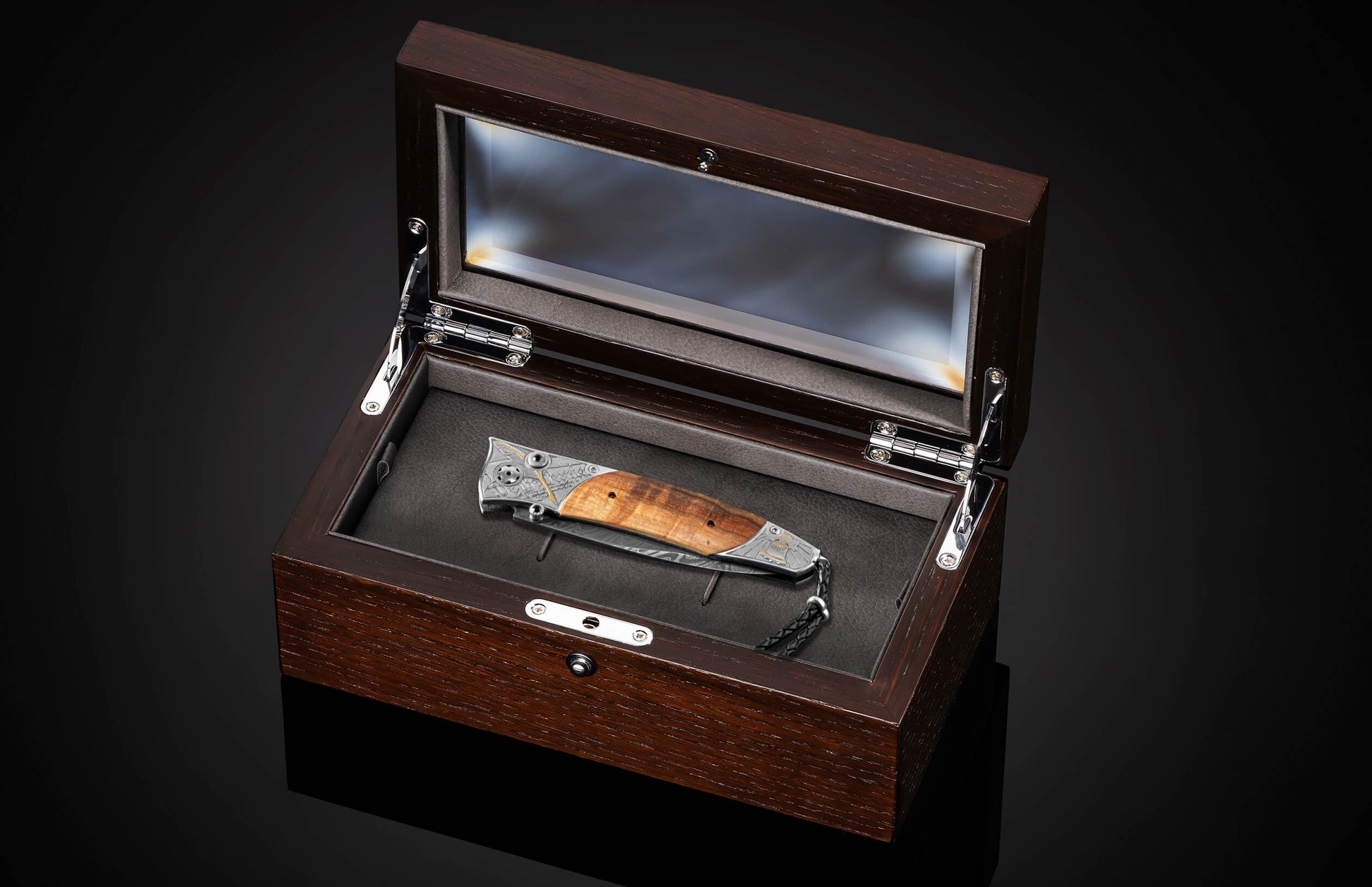 Folding knife with a patterned wooden handle and engraved metal accents, plus a black leather lanyard, in an open dark brown wooden box with glass lid.