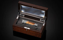 Folding knife with a patterned wooden handle and engraved metal accents, plus a black leather lanyard, in an open dark brown wooden box with glass lid.