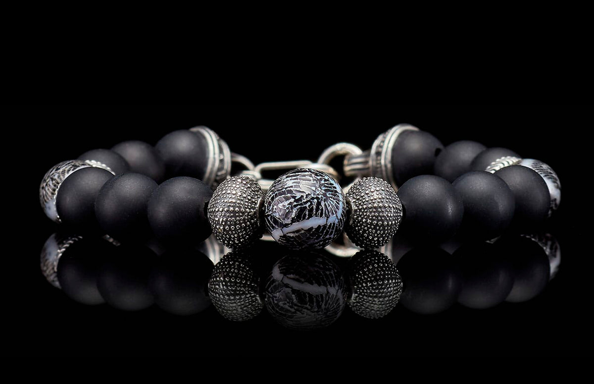 Black matte beaded bracelet with a central marbled bead and textured silver spacers, reflecting on a dark surface.