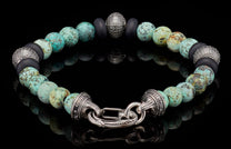 Beaded bracelet with mottled turquoise-colored and matte black beads, featuring silver studded accents and an ornate silver clasp, laid flat.