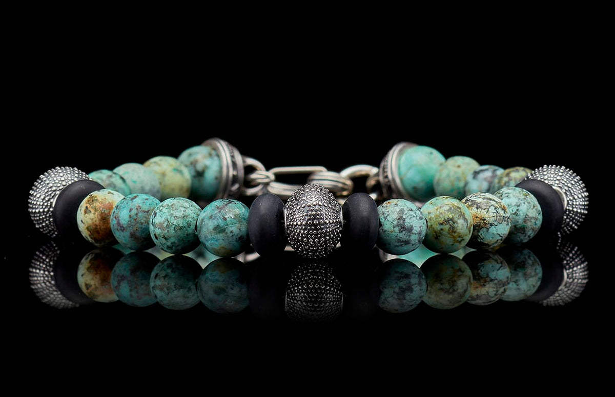 Beaded bracelet with mottled turquoise, matte black, and textured silver beads on a reflective surface.