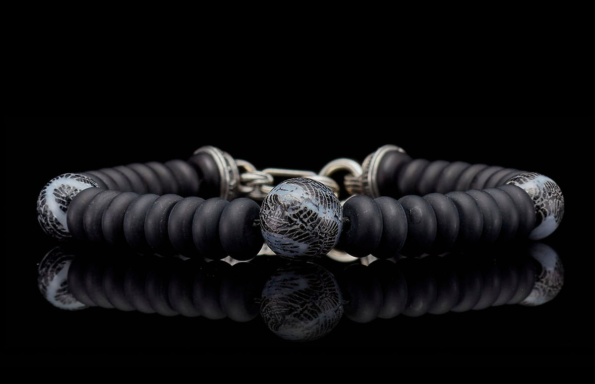 Black beaded bracelet with matte black and patterned grey beads, silver accents, reflecting on a dark surface.
