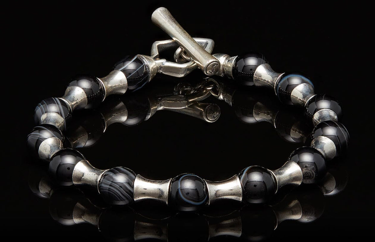 Silver and black beaded bracelet with a toggle clasp, featuring striped agate and smooth black beads separated by silver spacers on a reflective surface.