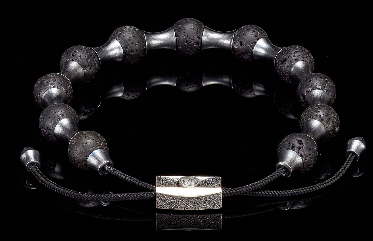 Black lava stone and silver-toned metal bracelet with textured beads and a rectangular patterned clasp on a reflective black surface.