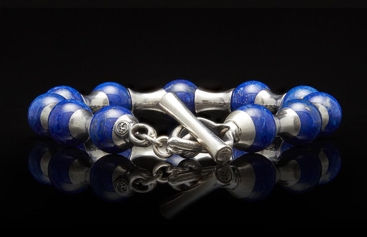 Blue lapis lazuli and silver beaded bracelet with a T-bar clasp, reflecting on a dark surface.