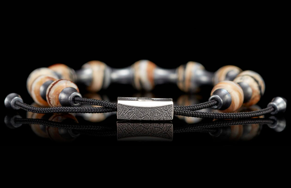 Braided black cord bracelet with alternating light brown patterned beads and metallic beads, featuring a textured rectangular silver clasp, reflected on a black surface.