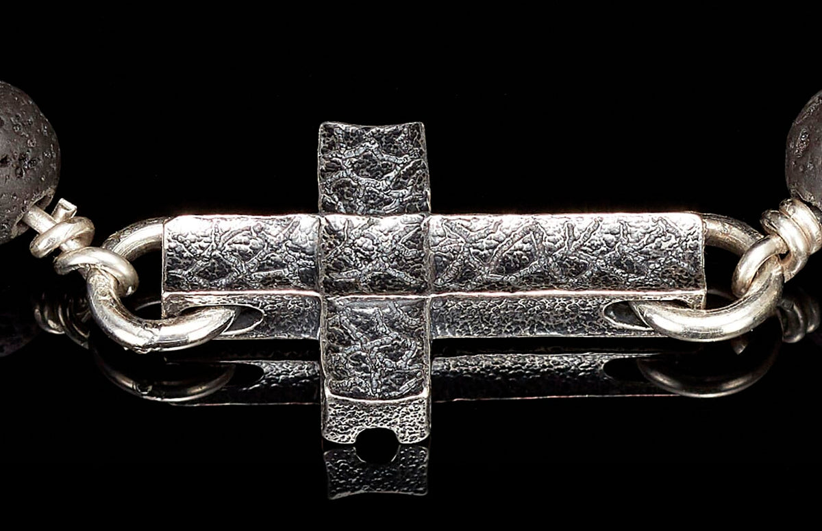 Cross-shaped silver jewelry clasp with textured surface, connected to a dark porous lava bead.