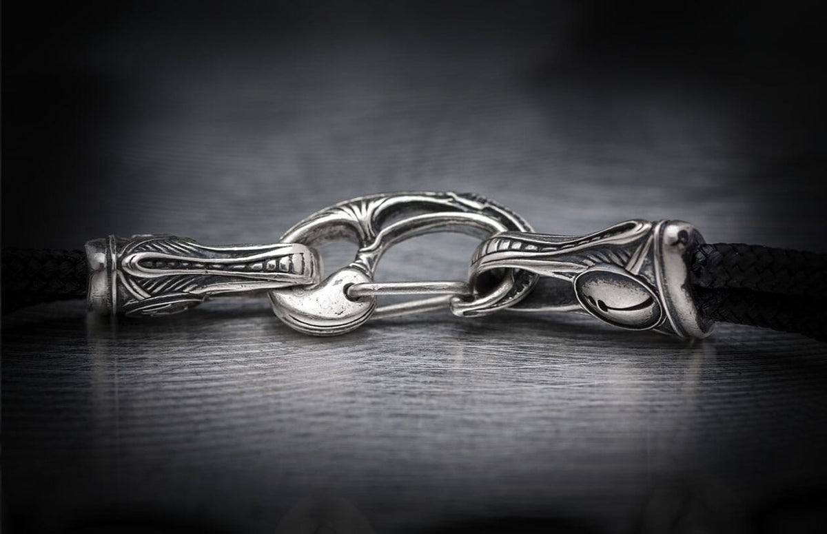 Silver jewelry clasp featuring elaborate decorative metalwork, connected to a black braided cord.