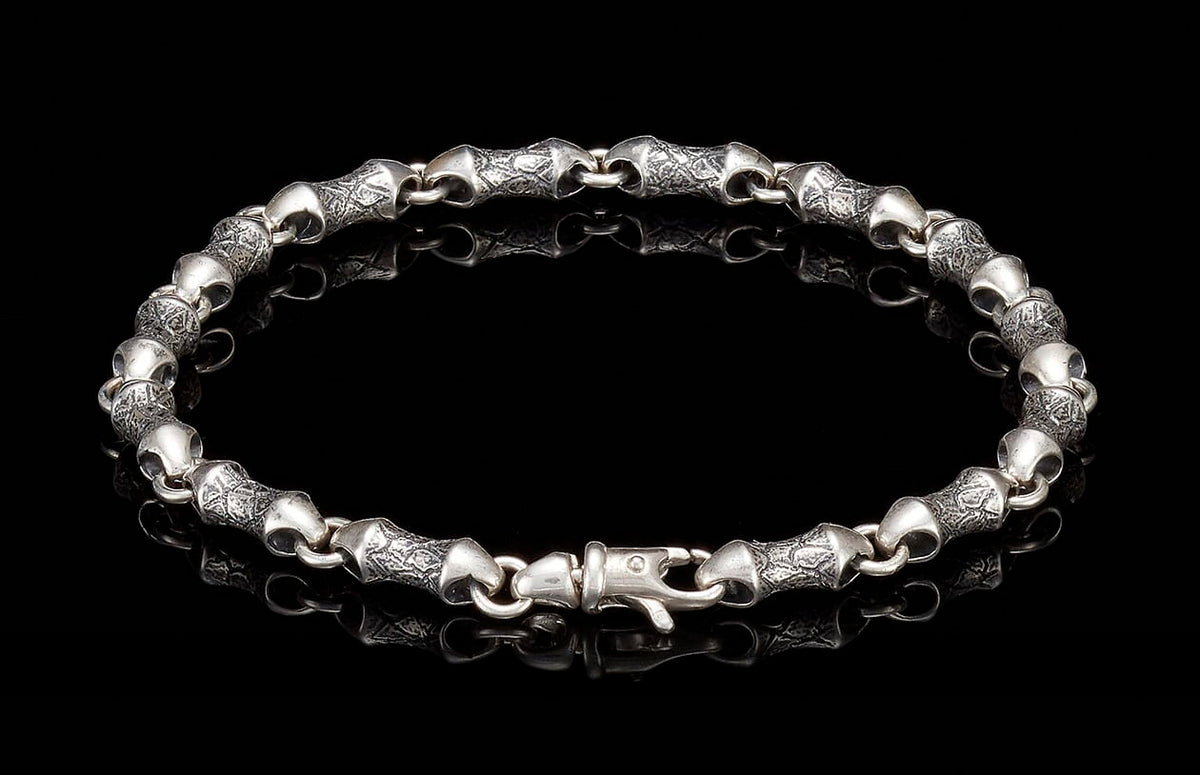 Silver bracelet with textured, barrel-shaped links and a decorative clasp on a reflective black surface.
