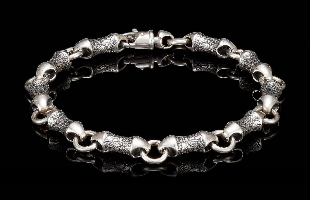 Textured silver bracelet featuring stylized, elongated links with an engraved pattern and a toggle clasp, laid flat.