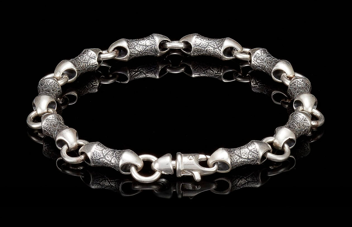Silver-toned metal bracelet with textured, bamboo-shaped links and a lobster clasp, reflecting on a black surface.