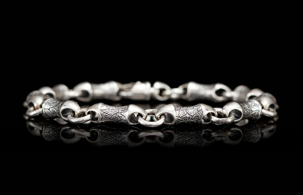 Silver metal bracelet with patterned and smooth bone-shaped links reflected on a black surface.