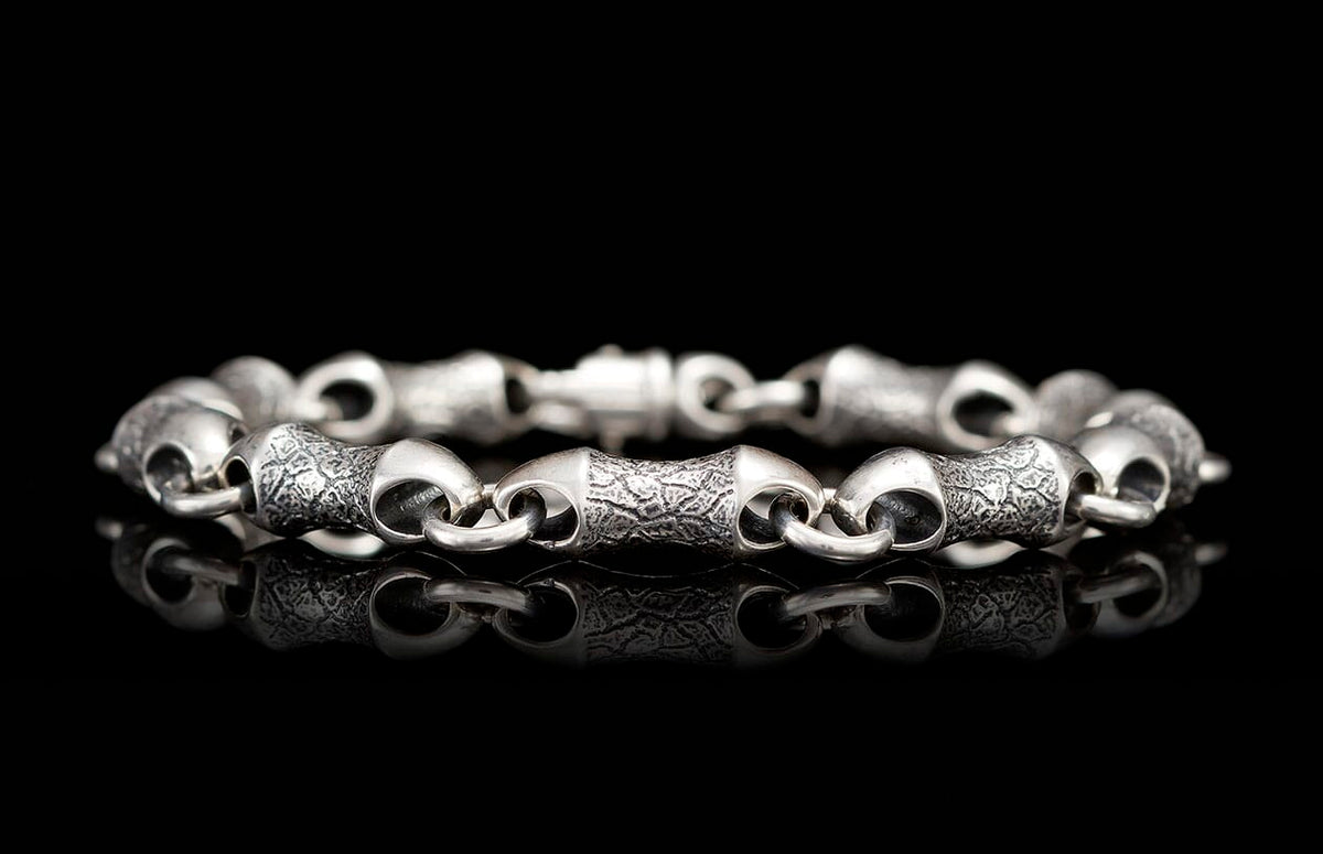 Chunky silver bracelet with alternating textured and smooth barrel-shaped links, on a reflective black surface.