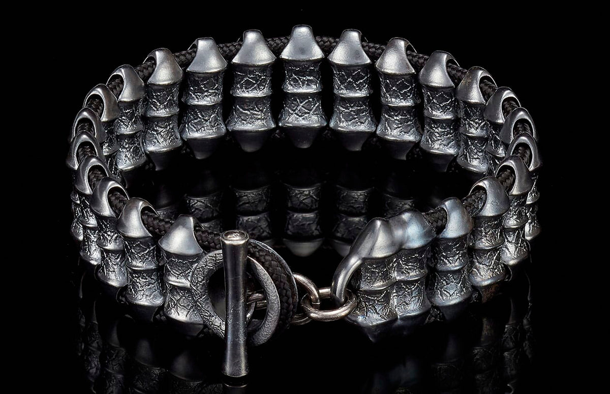 Dark silver metal bracelet with textured cylindrical beads, black braided cord, and a toggle clasp on a reflective surface.