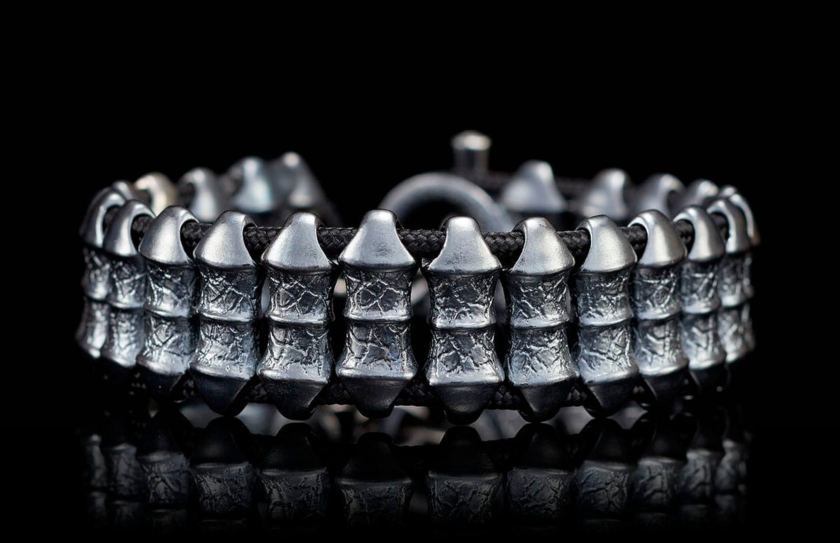 Dark silver metal bracelet with textured rectangular beads on a braided black cord, on a reflective surface.