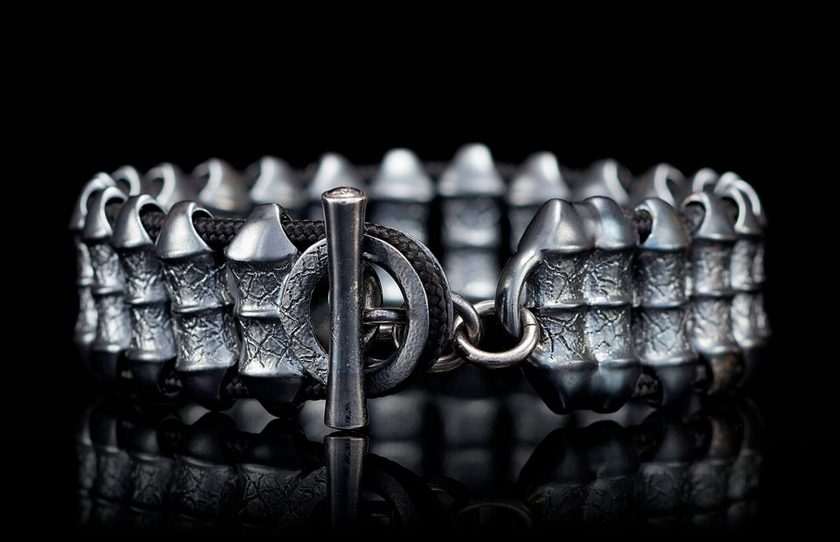 Dark silver bracelet with rugged textured metal segments and black cord, featuring a toggle clasp.