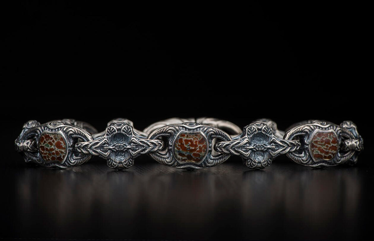 Oxidized silver bracelet with ornate scrollwork links and reddish-brown, green-veined patterned stones, on a dark surface.