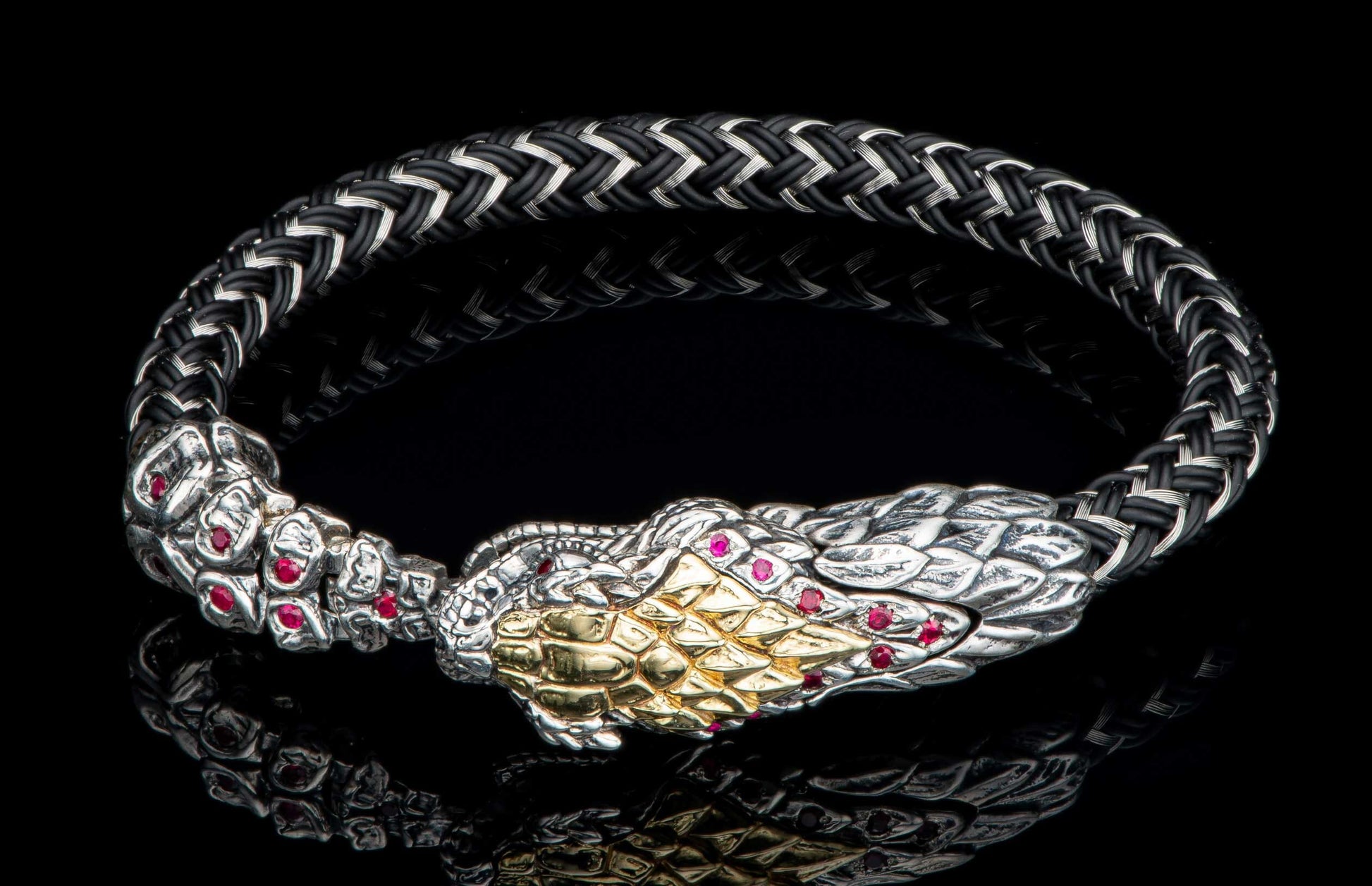 Black braided bracelet with a silver snake head featuring gold scales and red ruby accents, reflecting on a dark surface.