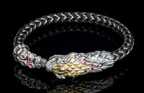 Black braided bracelet with a silver snake head featuring gold scales and red ruby accents, reflecting on a dark surface.