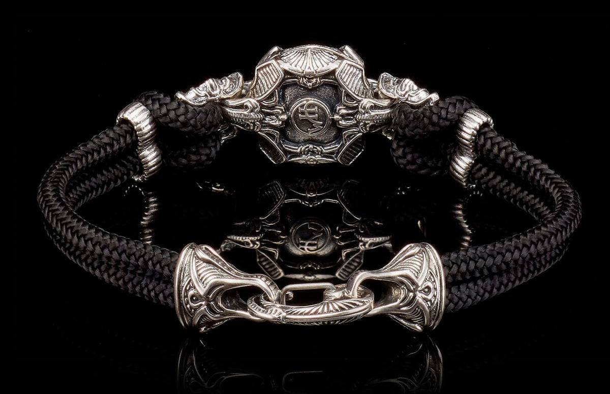 Black braided cord bracelet featuring an ornate silver-toned stylized helmet centerpiece and decorative hook clasp.