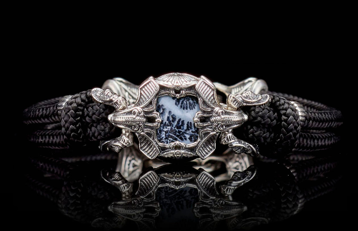 Ornate silver-toned bracelet with a black braided band and a central black and white dendritic stone framed by stylized dragon heads.