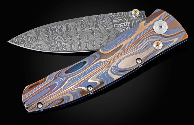 Handcrafted pocketknife from William Henry features a light and resilient titanium frame that is laser-engraved in swirls of color. The blade is hand-forged damascus steel. The button lock and thumb stud are inset with citrine gemstones.