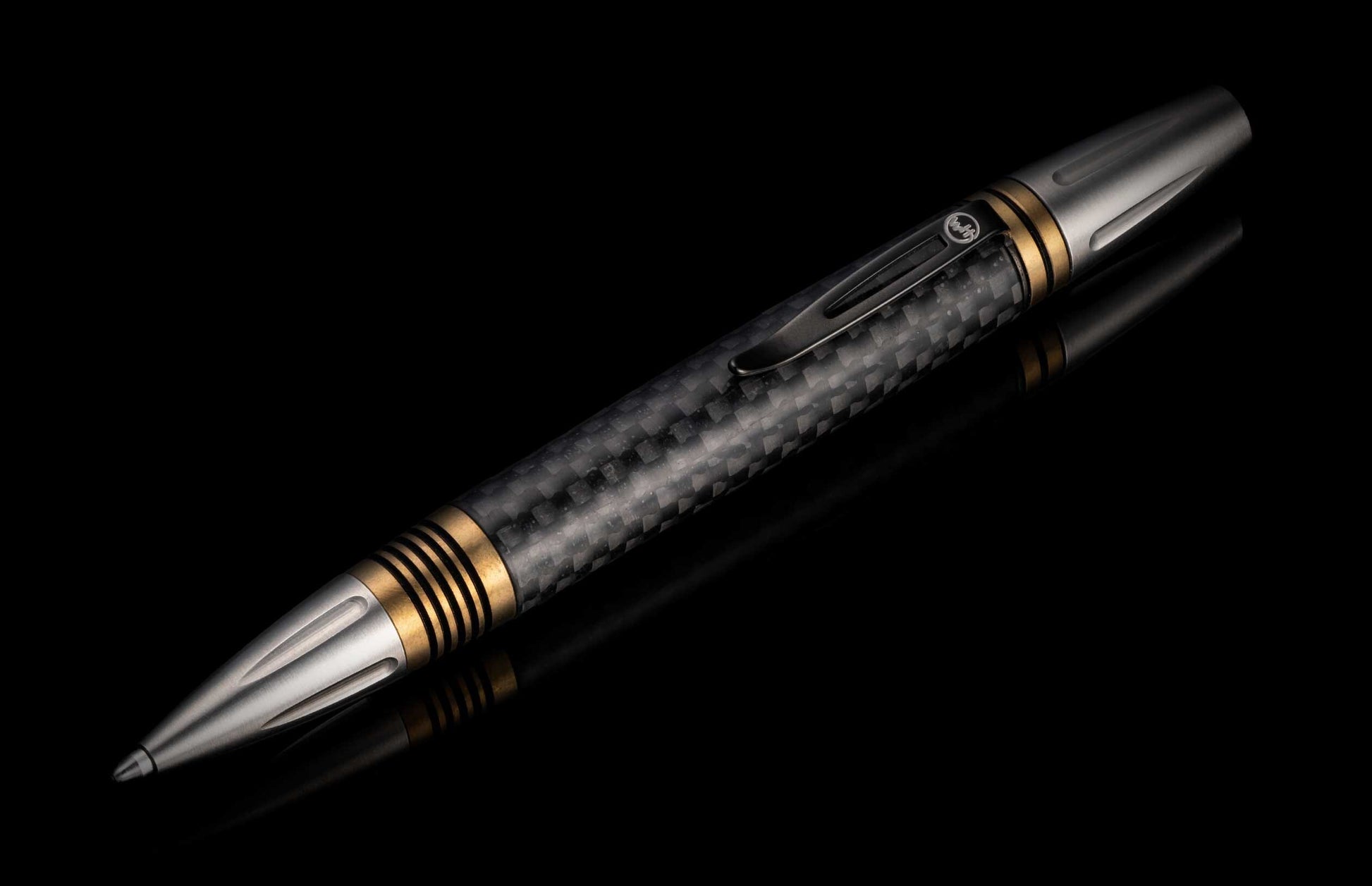 A classic executive pen from William Henry. The distinctive carbon fiber barrel is flanked by brass spacers with deep grooves, and the tip and top section are crafted from stainless steel with art deco details. The pen is displayed on a black background.