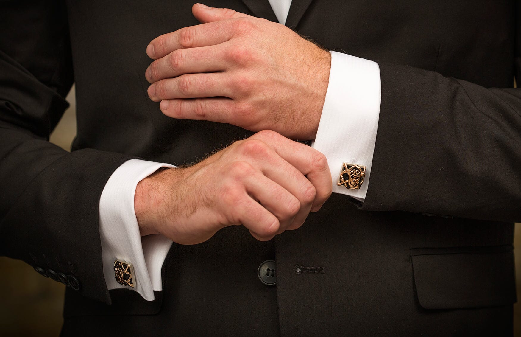 Square bronze cufflinks with an intricate, stylized openwork design, worn on white shirt cuffs with a black suit.