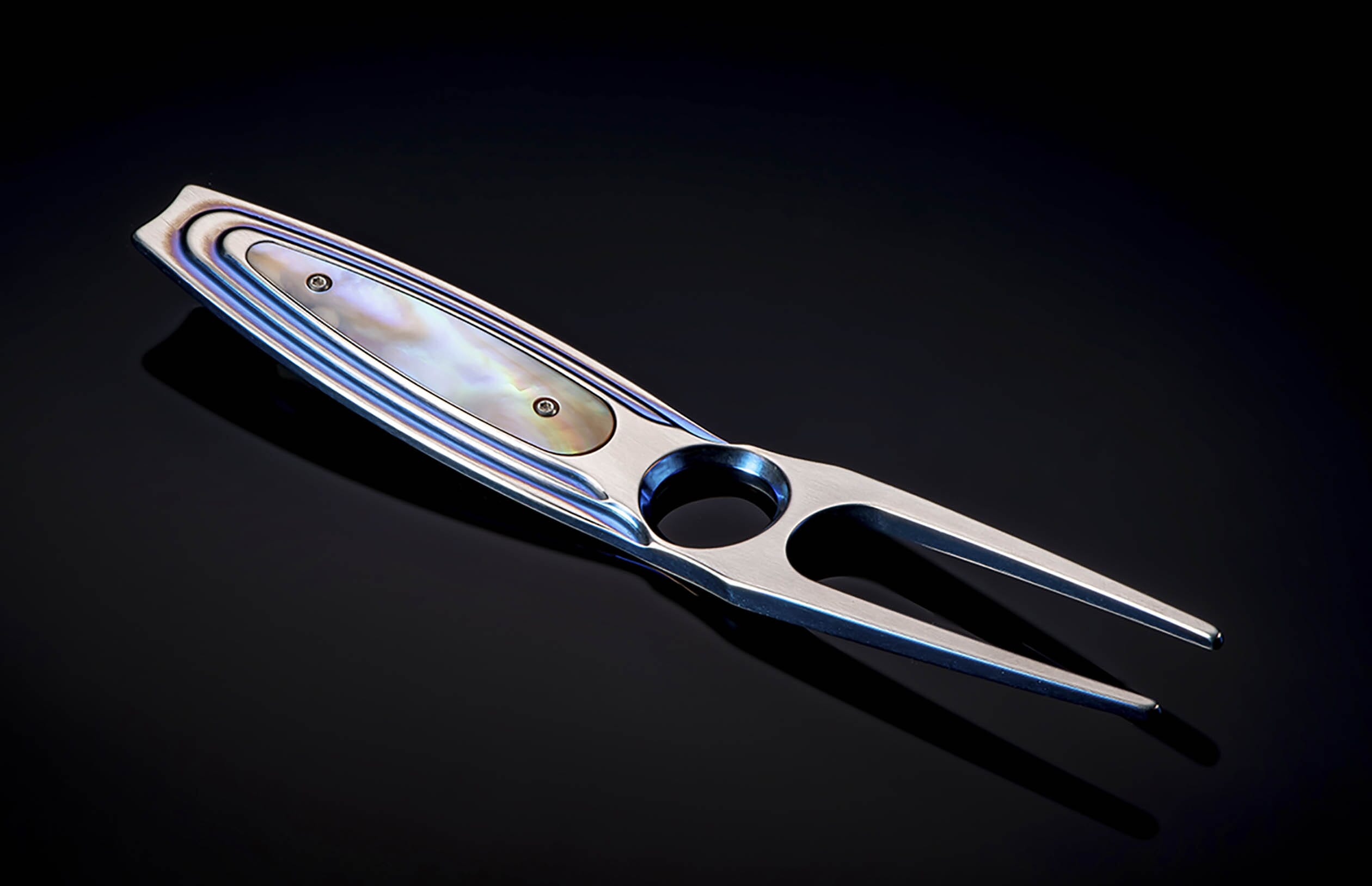 Iridescent blue metal golf divot tool with mother-of-pearl inlay, angled on a dark reflective surface.