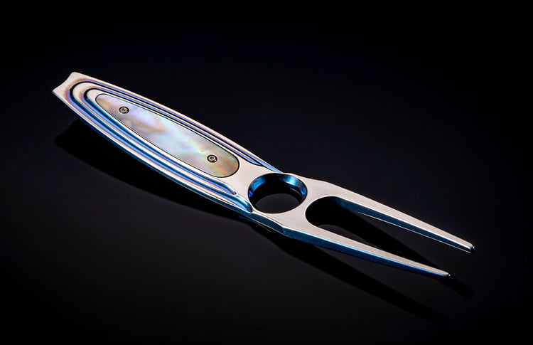 Iridescent blue metal golf divot tool with mother-of-pearl inlay, angled on a dark reflective surface.