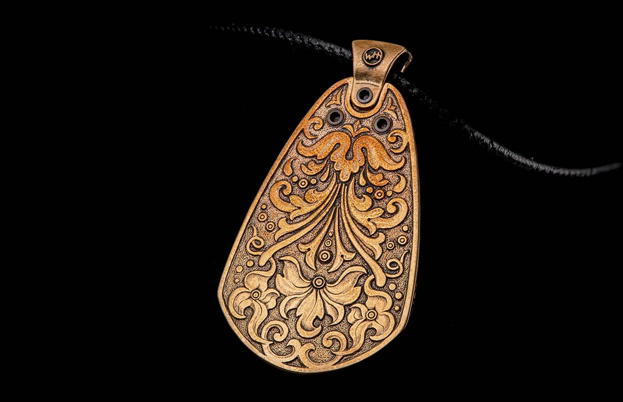 Bronze pendant with intricate floral and swirling decorative motifs, hanging from a black braided cord on a black background.