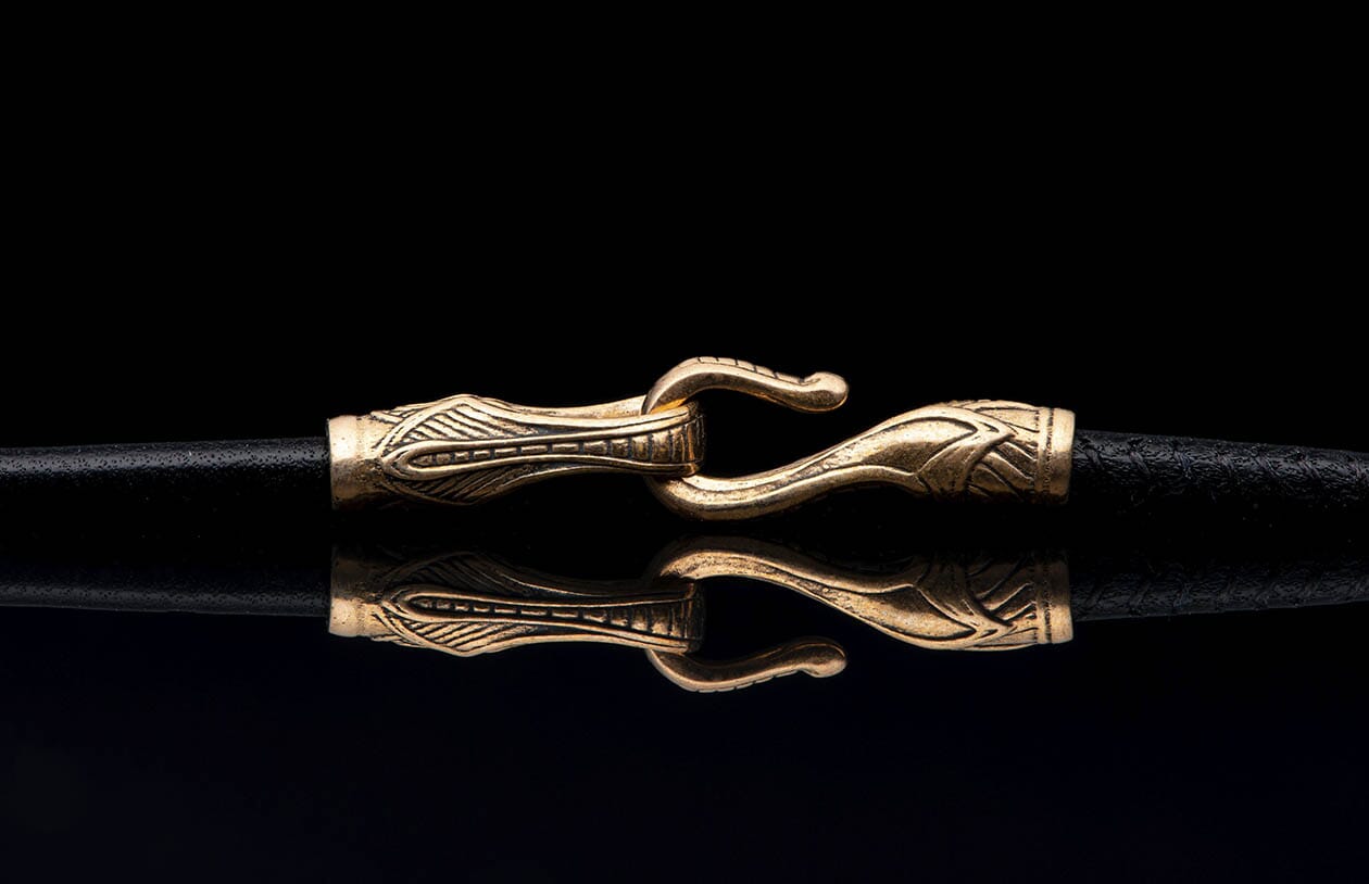 A piece of jewelry featuring a decorative bronze clasp with an ornate pattern on black textured cords, reflected below.