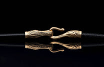 A piece of jewelry featuring a decorative bronze clasp with an ornate pattern on black textured cords, reflected below.