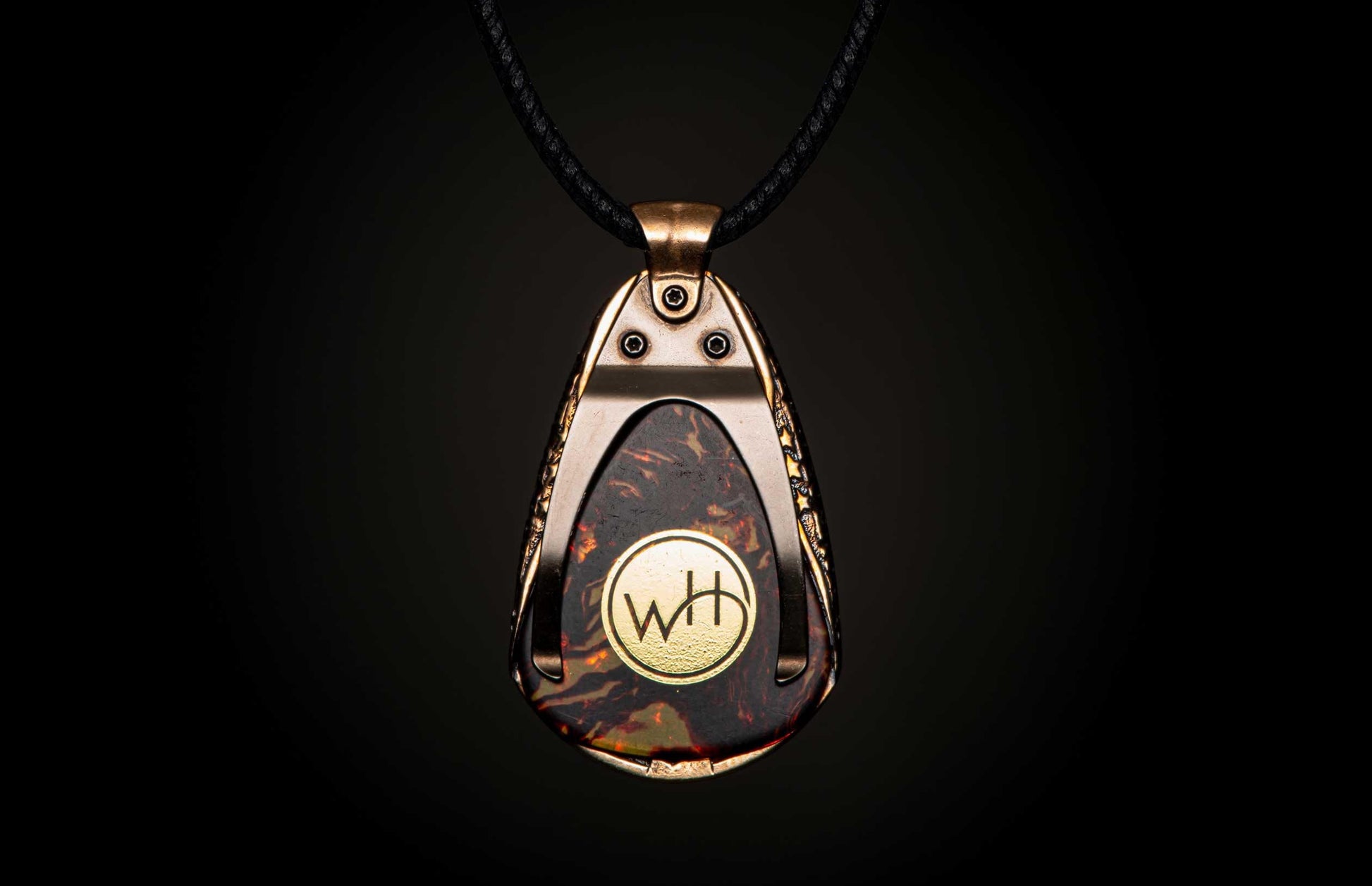 Dark mottled pendant with a bronze-toned frame, featuring a gold 'WH' emblem, on a black braided cord.