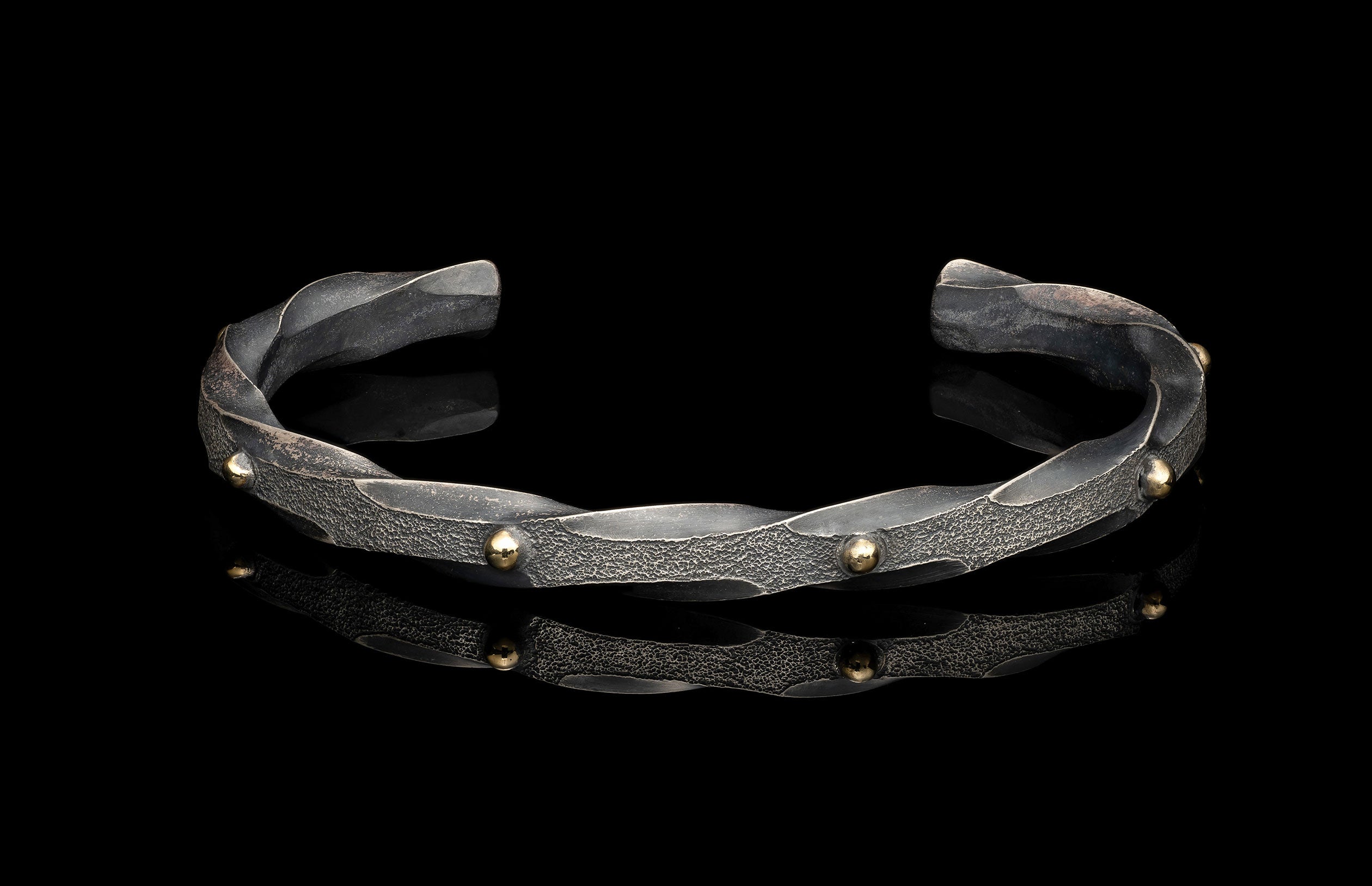 Textured silver cuff bracelet with twisted design and golden spherical studs, reflecting on a black surface.