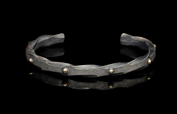Textured silver cuff bracelet with twisted design and golden spherical studs, reflecting on a black surface.
